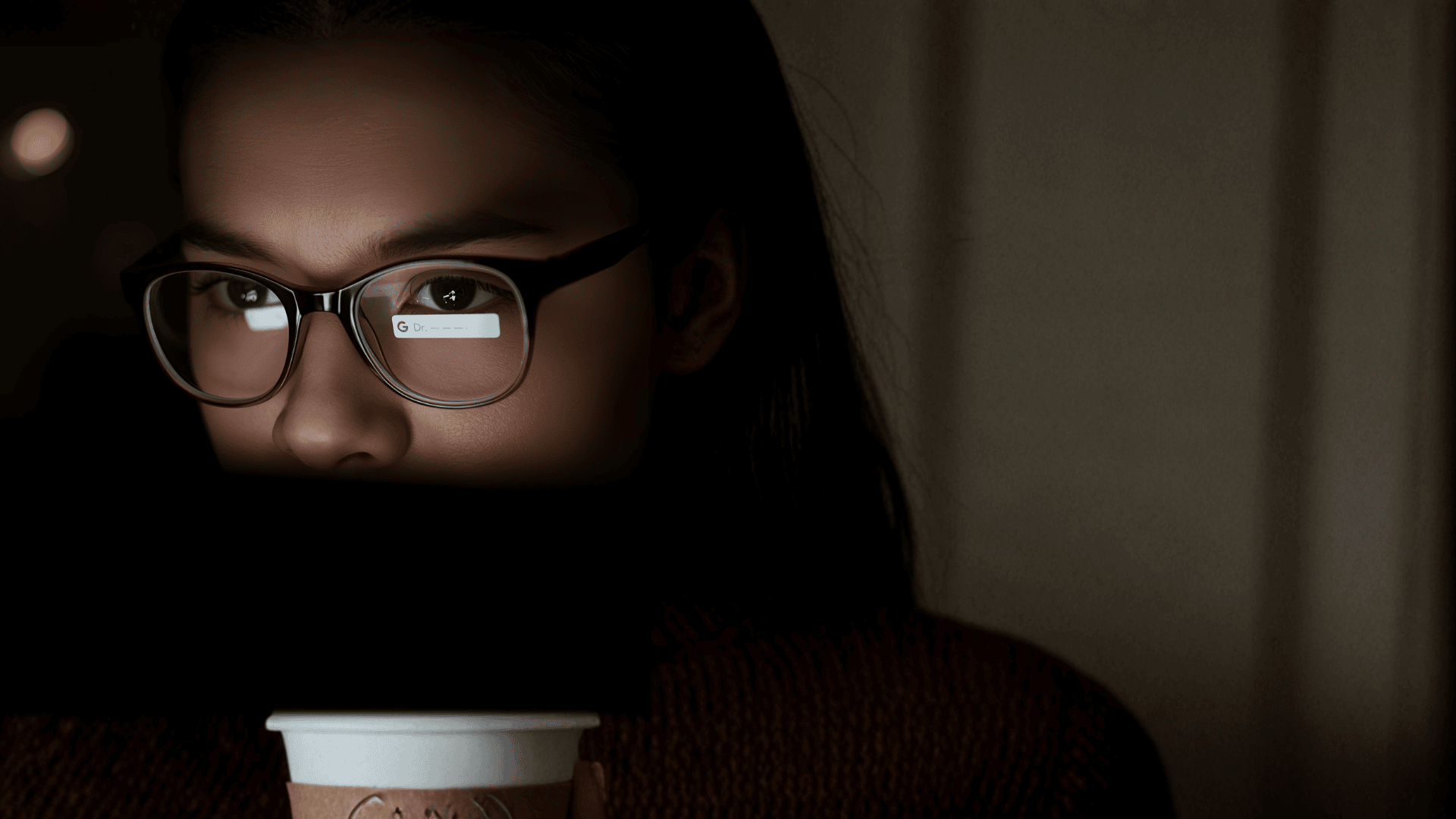 Γυναίκα με γυαλιά βλέπει οθόνη laptop ενώ κρατά κούπα καφέ, σε σκοτεινό δωμάτιο.